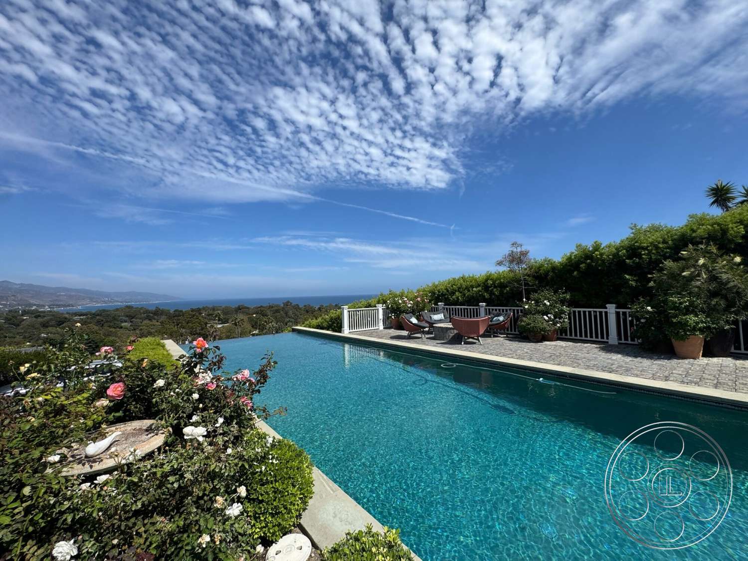 Americana 113 - exterior, ocean view, white picket fence, infinity pool, suburban backyard, wooden lounge chairs, stone pool deck, brick patio flooring, blue sky with clouds, concrete coping, landscaped garden with roses