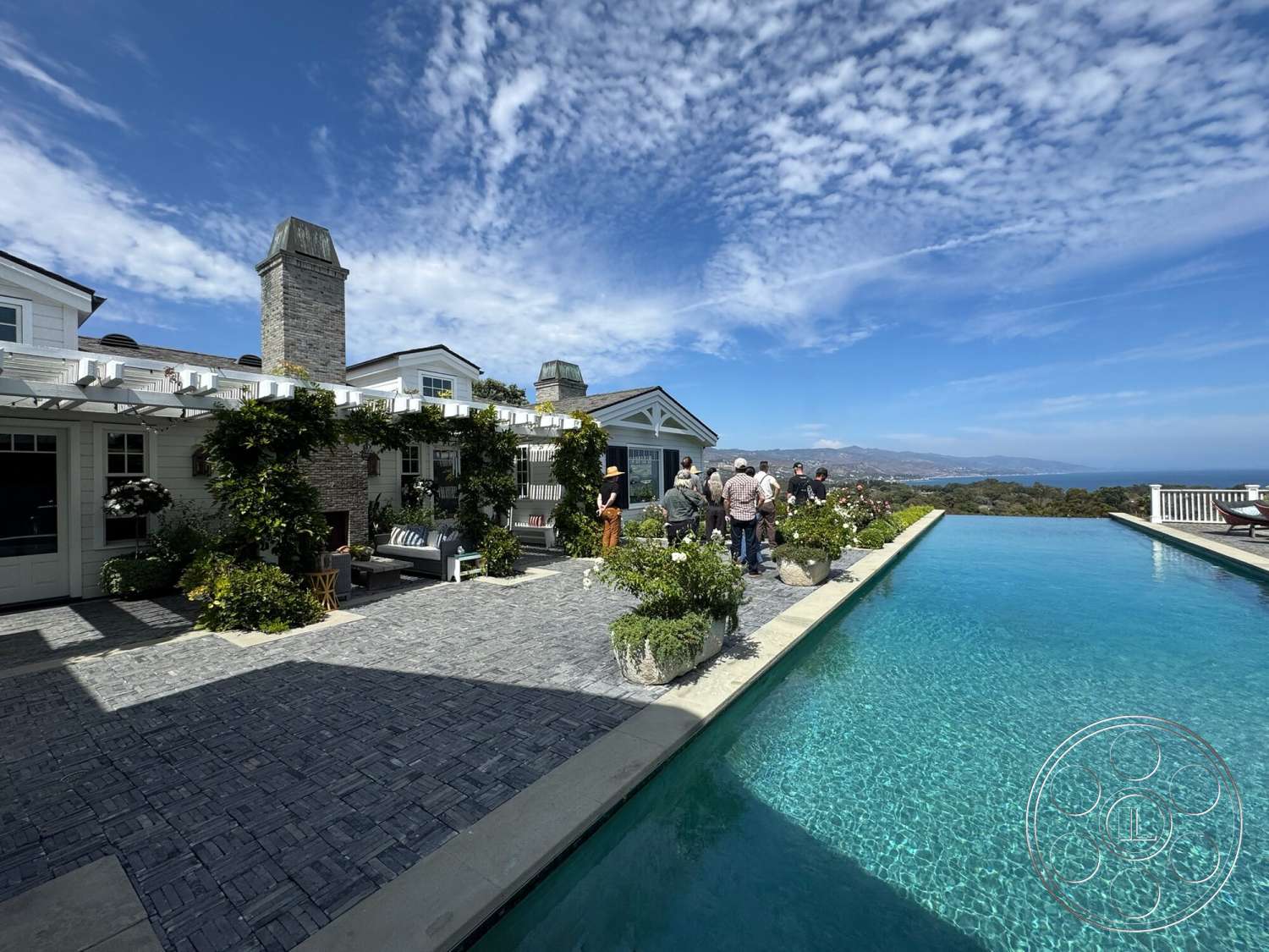 Americana 113 - exterior, glass sliding doors, city skyline view, infinity pool, wooden outdoor furniture, stone chimney, concrete pool border, paver patio flooring, landscaped garden with flowering plants, pergola with climbing plants, modern home with gable roof, deck with lounge chairs, partial mountain view