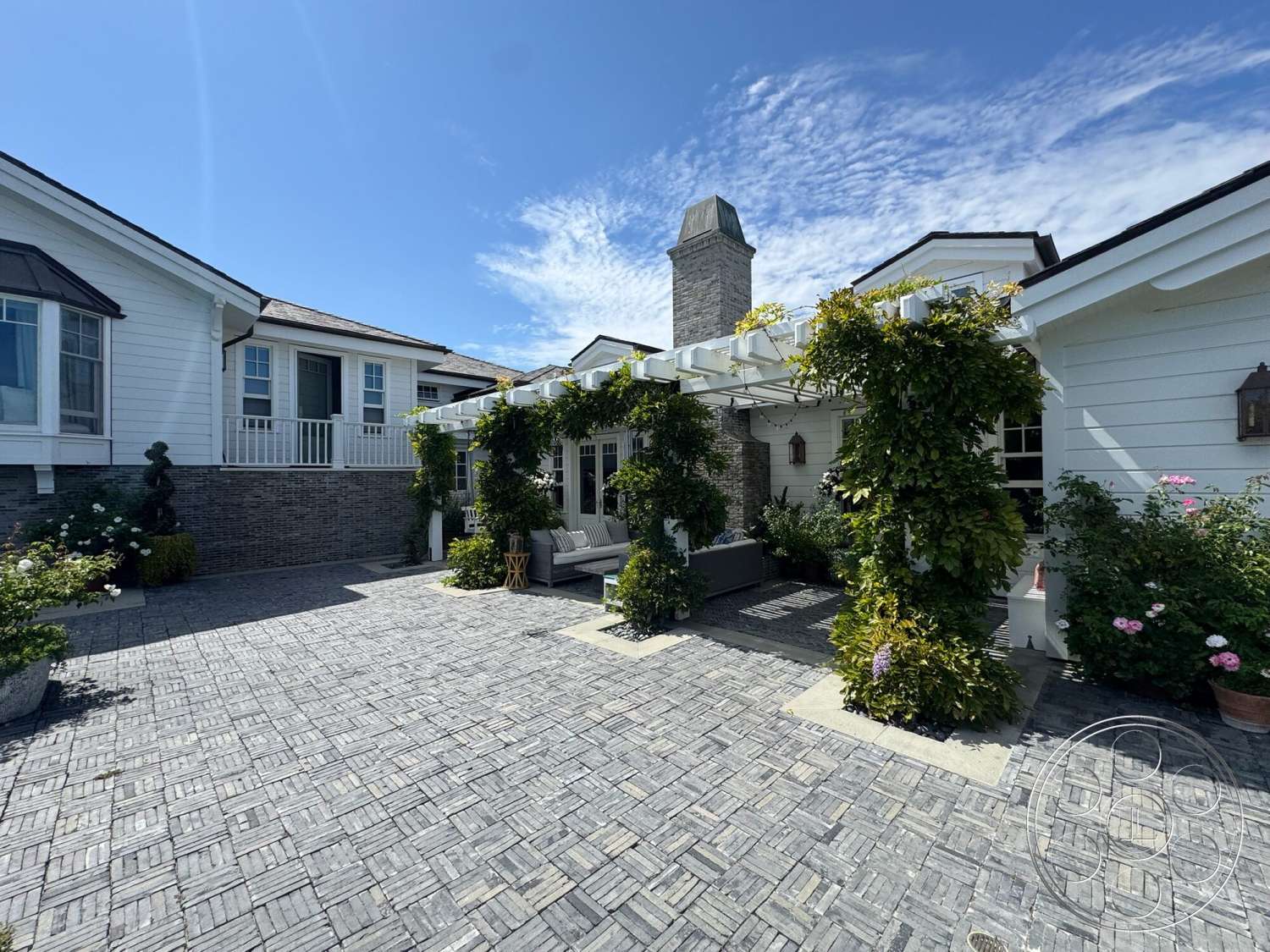 Americana 113 - exterior, landscaped garden, outdoor seating area, gabled roof, metal light fixtures, vertical siding, stone chimney, wood pergola, white painted walls, horizontal wood railing, raised flower beds, gray brick pavers driveway