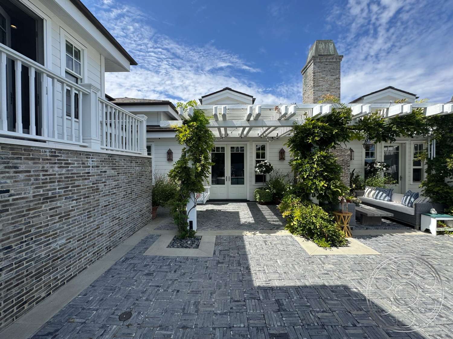 Americana 113 - natural light, exterior, paver driveway, outdoor seating area, stone chimney, two-story home, white painted walls, lush greenery landscaping, slate patio flooring, vertical planters, wooden balcony railing, concrete border, pergola structure, bricks exterior wall