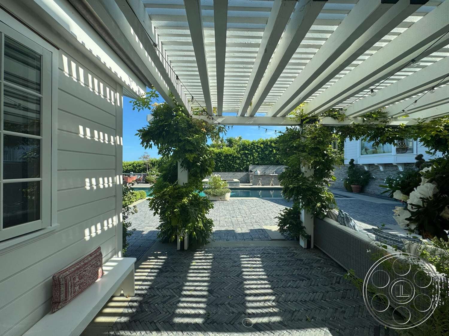 Americana 113 - exterior, rectangular swimming pool, white wood siding, concrete pool deck, sunny outdoor setting, outdoor seating area with cushions, landscaped garden with greenery, wooden pergola with slatted roof, green climbing plants around pergola, herringbone paver patio flooring, stone wall surrounding pool area, view of pool from patio