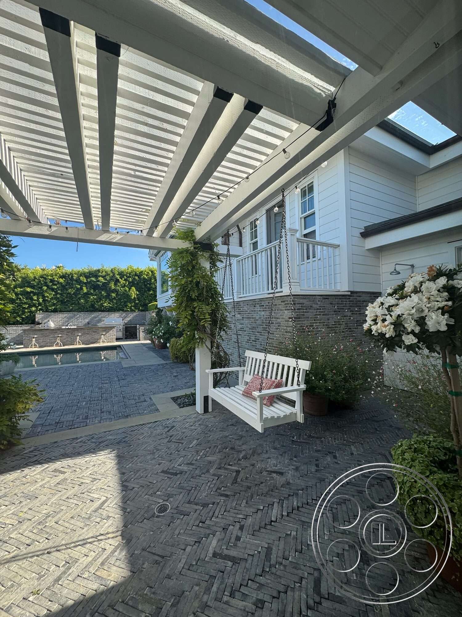 Americana 113 - exterior, surrounding greenery, outdoor living space, brick accent wall, rectangular pool, sunny outdoor setting, textured stone pavers, herringbone pattern stone patio flooring, wooden pergola with white slats, built-in swinging bench with white finish, green landscaping with potted plants