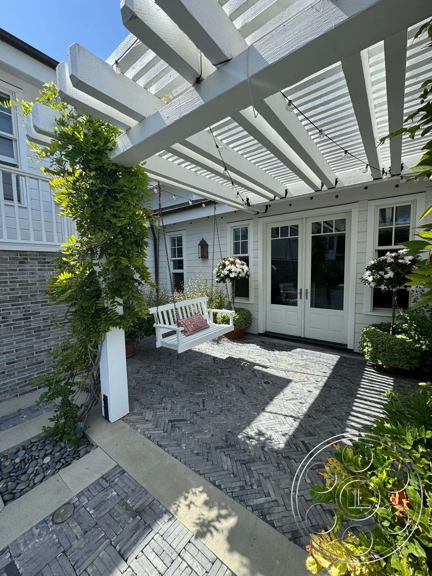 Americana 113 - exterior, potted plants, landscaped garden area, glass double doors, hanging lantern light fixture, herringbone brick patio flooring, white wooden pergola, white swing bench with cushion, stone border edges, green climbing plants