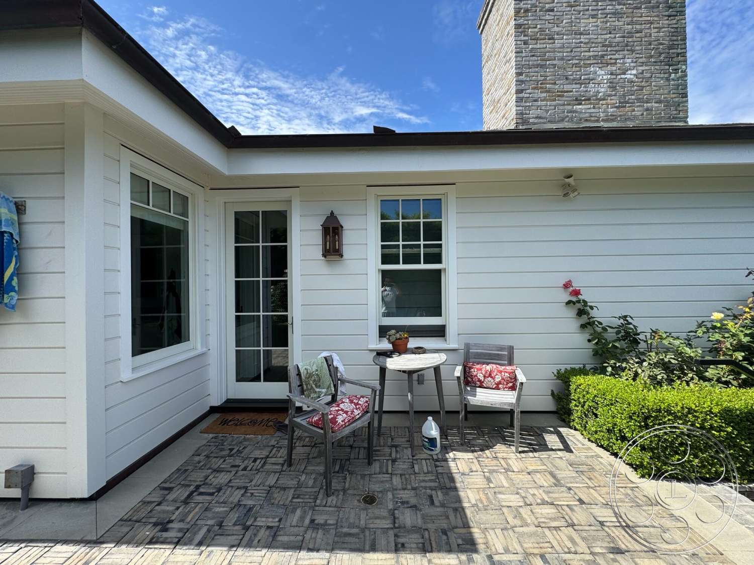 Americana 113 - exterior, landscaped garden, potted plants, wooden patio furniture, welcome mat, round outdoor table, vertical wood siding, boxwood hedge, brick chimney, lantern-style outdoor lighting, concrete patio flooring, floral outdoor cushions, window with grids