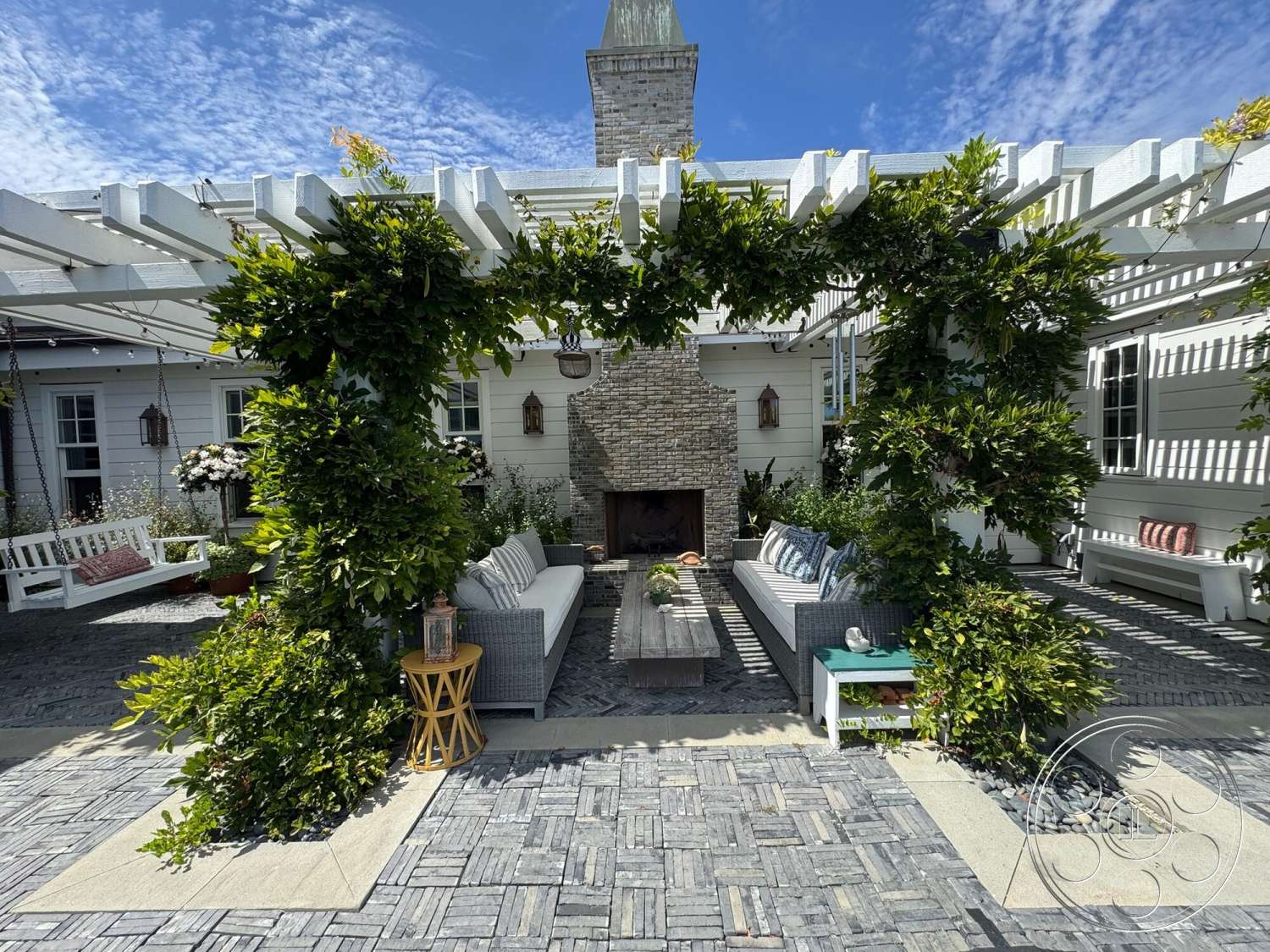 Americana 113 - exterior, outdoor seating area, woven outdoor furniture, integrated landscaping, wood beam pergola, stone fireplace surround, white painted siding, natural stone accent wall, decorative outdoor table, gray stone patio flooring, checkered pavers patio, green climbing vines, hanging lantern outdoor lighting, potted plants surrounding seating area