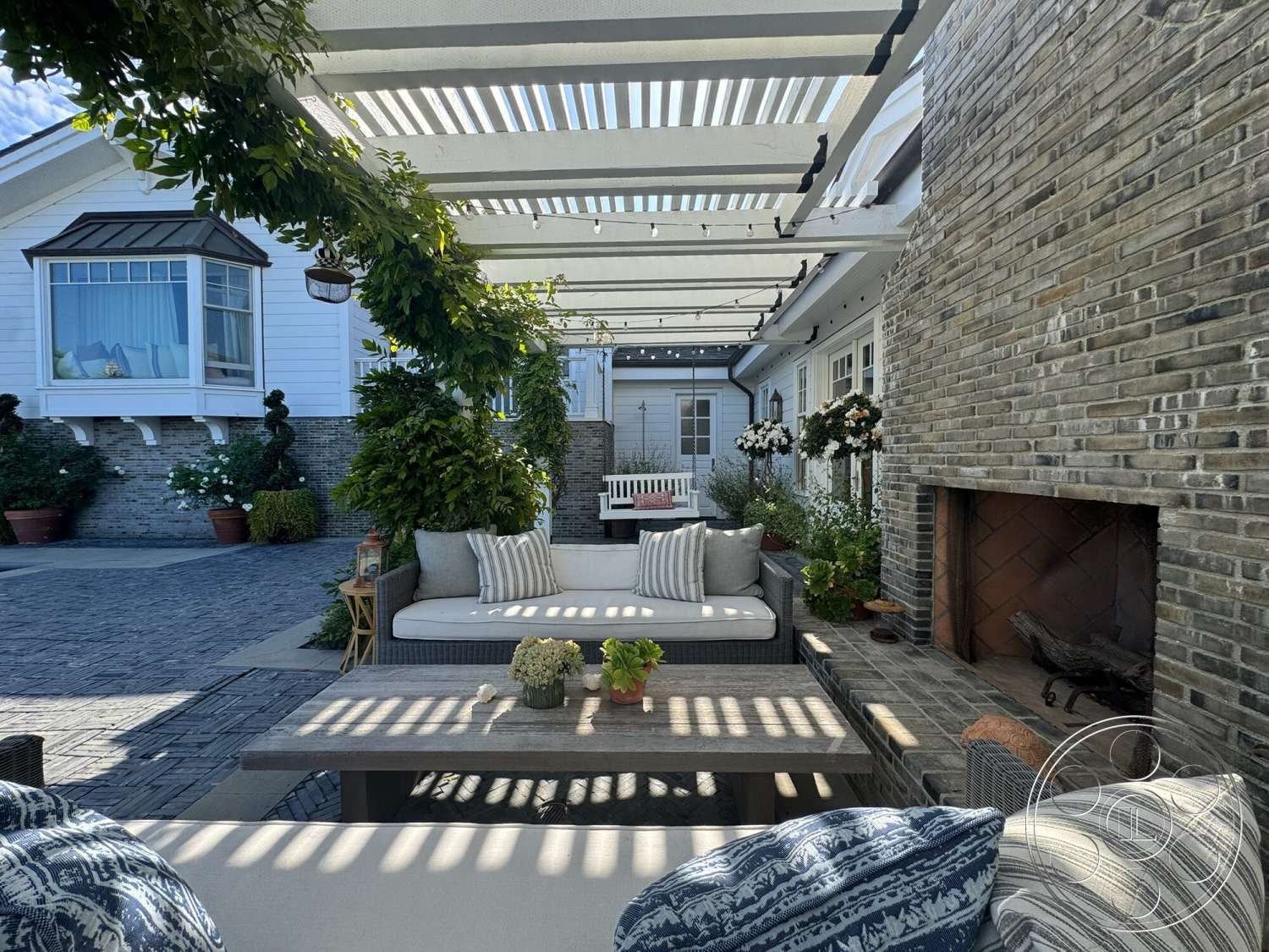 Americana 113 - exterior, landscaped garden, wood coffee table, outdoor dining area, striped outdoor cushions, gray brick walls, wood beam pergola, flower planters, brick outdoor fireplace, paver stone flooring, modern patio seating area, gray wicker outdoor furniture, sunlit courtyard setting