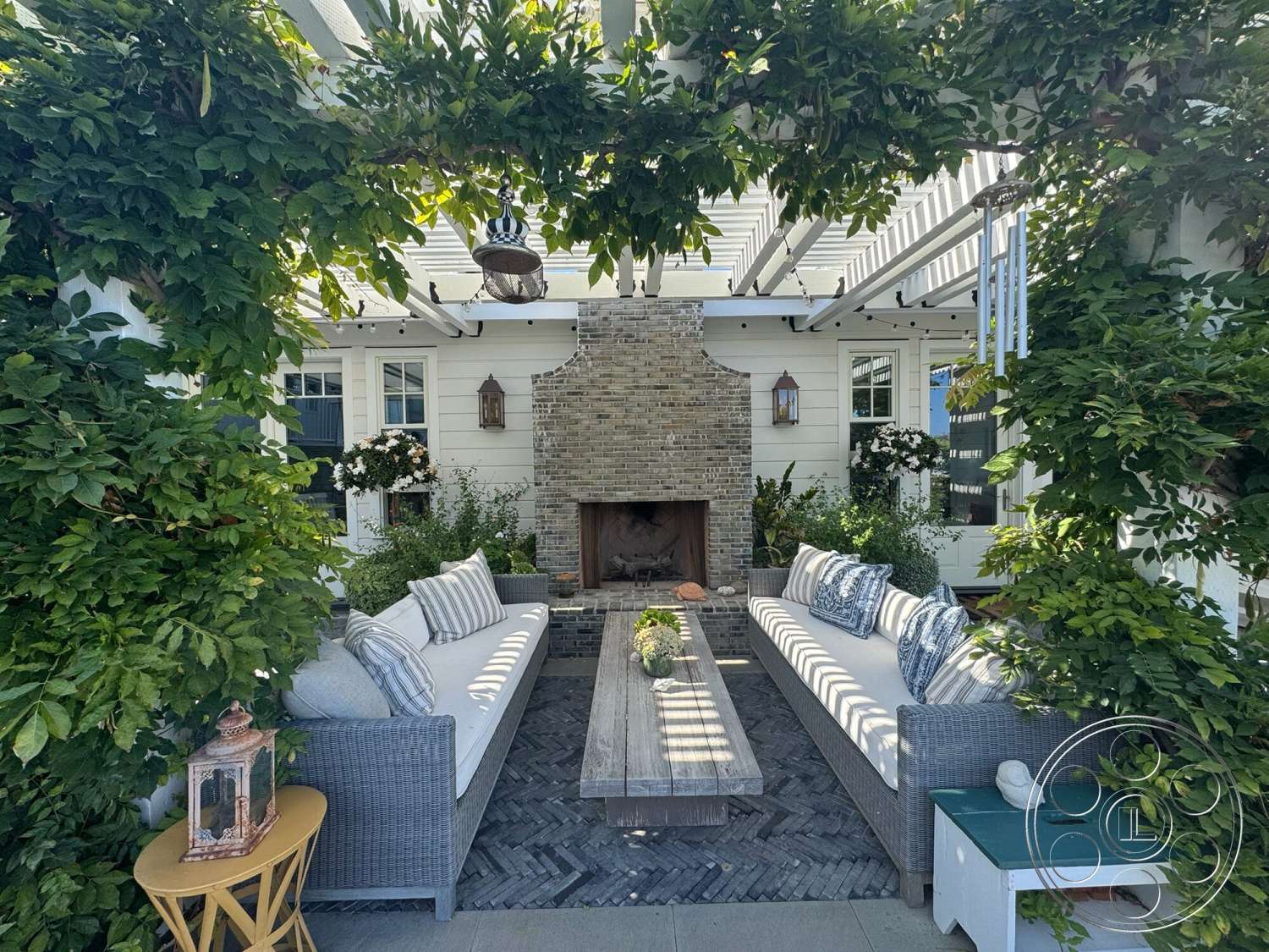 Americana 113 - exterior, woven outdoor furniture, decorative lanterns, rectangular coffee table, striped outdoor cushions, brick fireplace wall, gray stone patio flooring, wooden beam pergola, lush landscaping with climbing vines, textured plant arrangements