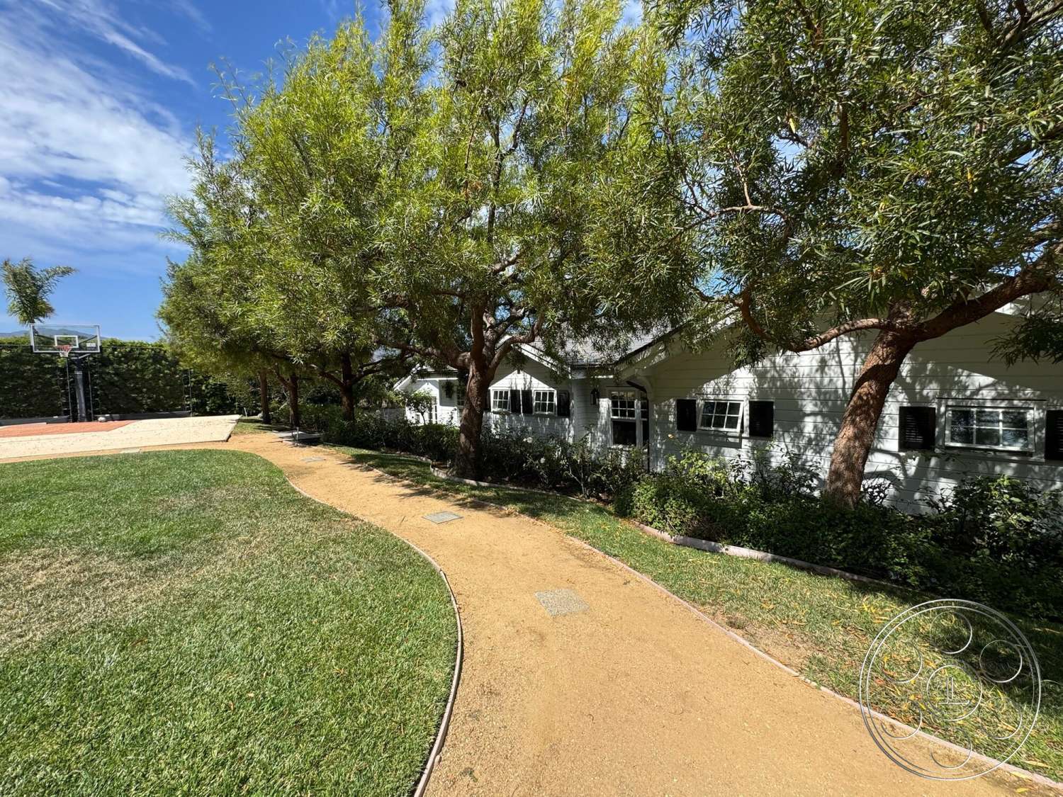 Americana 113 - exterior, landscaped garden, flat roof, lush green grass, outdoor basketball court, black shutters, shade trees, urban backyard, brick patio border, white painted walls, cement driveway, curved gravel pathway