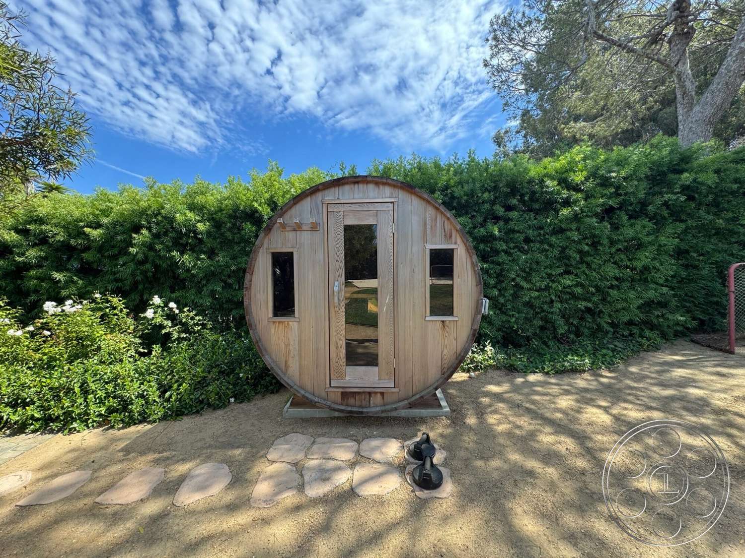 Americana 113 - rustic design elements, natural stone pathway, landscaped garden setting, wooden barrel sauna exterior, sunny outdoor environment, glass door with wooden frame, circular sauna structure, small windows with wooden trim, green hedge surrounding, ground-level placement