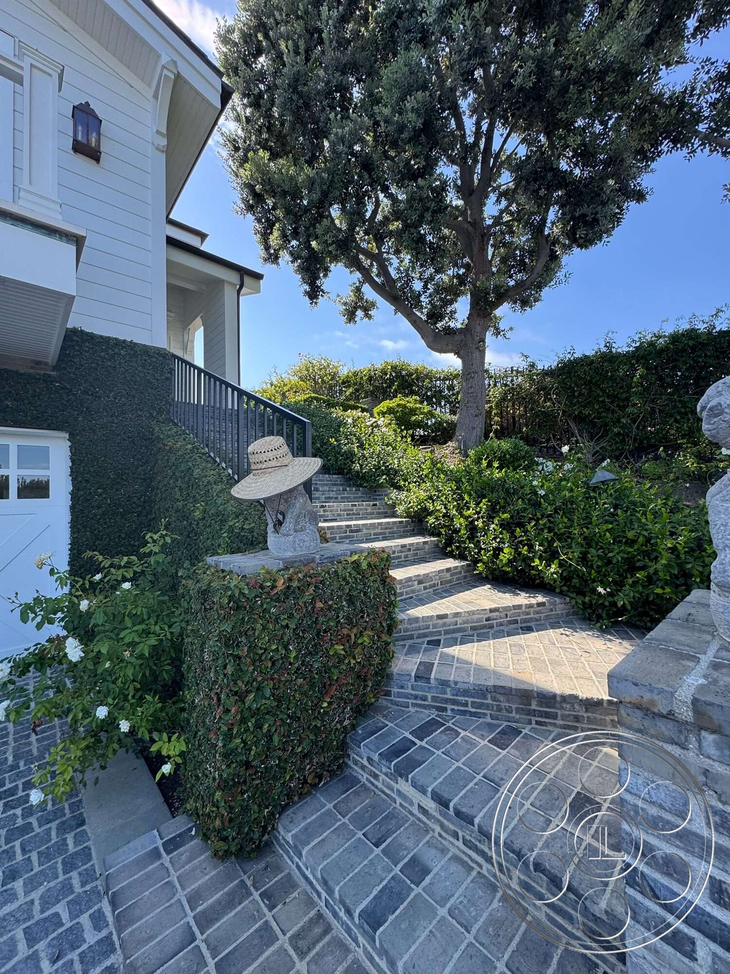 Americana 113 - exterior, landscaped garden, green foliage, cobblestone pathway, trimmed hedges, wooden railings, stone steps, sunny outdoor setting, brick retaining wall, decorative stone features, curved steps, mature tree, white flowering plants