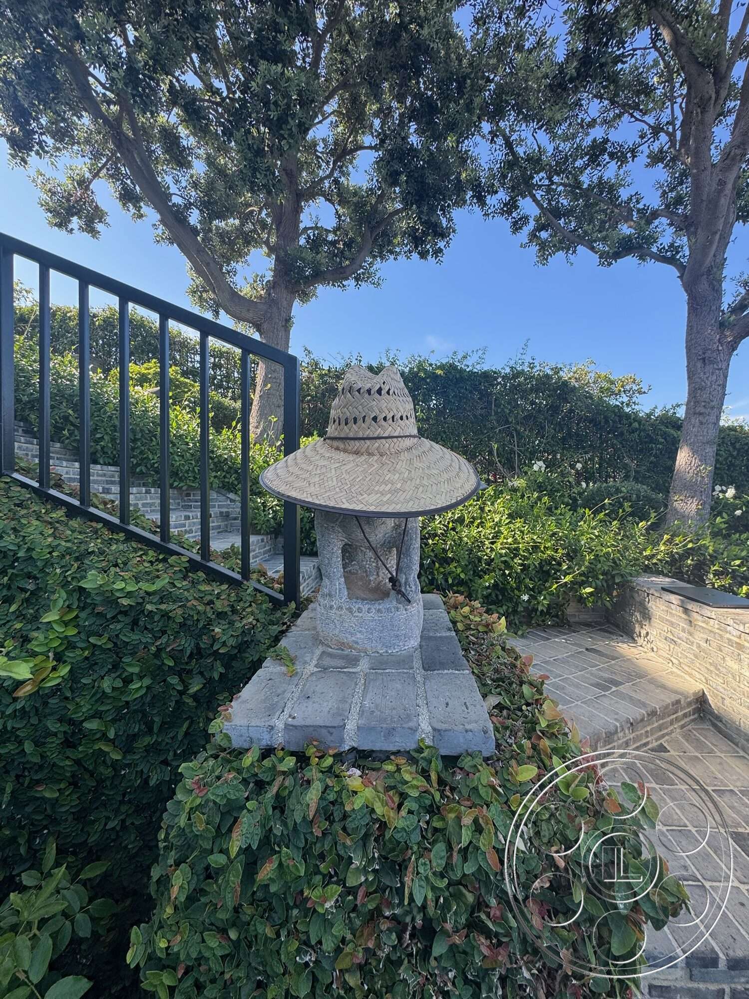 Americana 113 - exterior, green landscaping, metal railing, concrete pathway, tall trees, lush hedges, stone pedestal base, woven straw hat