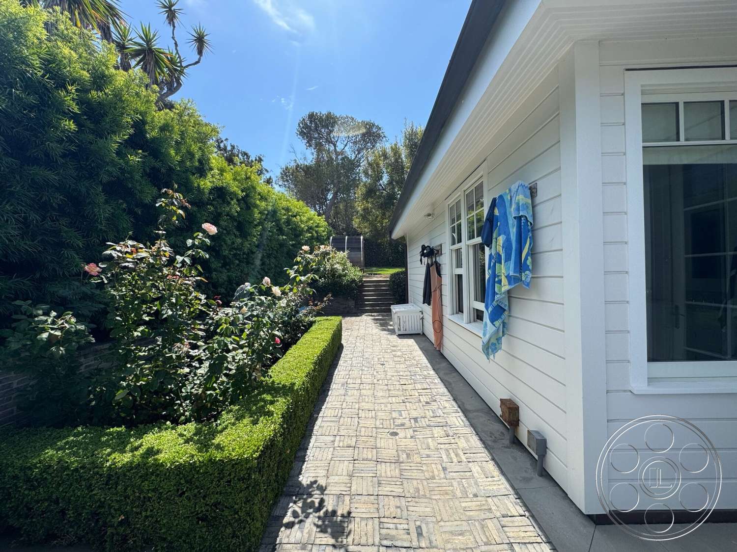 Americana 113 - exterior, concrete steps, lush green hedges, rose garden, sunny outdoor setting, landscaped garden area, herringbone patterned stone walkway, white horizontal lap siding walls, towel hanging on wall