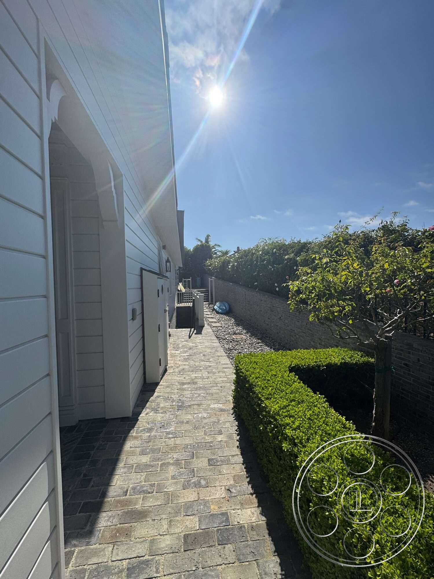 Americana 113 - exterior, lush greenery, concrete block walls, brick walkway, shadow play, hedge landscaping, sunny outdoor setting, paved garden path, white horizontal siding, light gray facade, structured pathway
