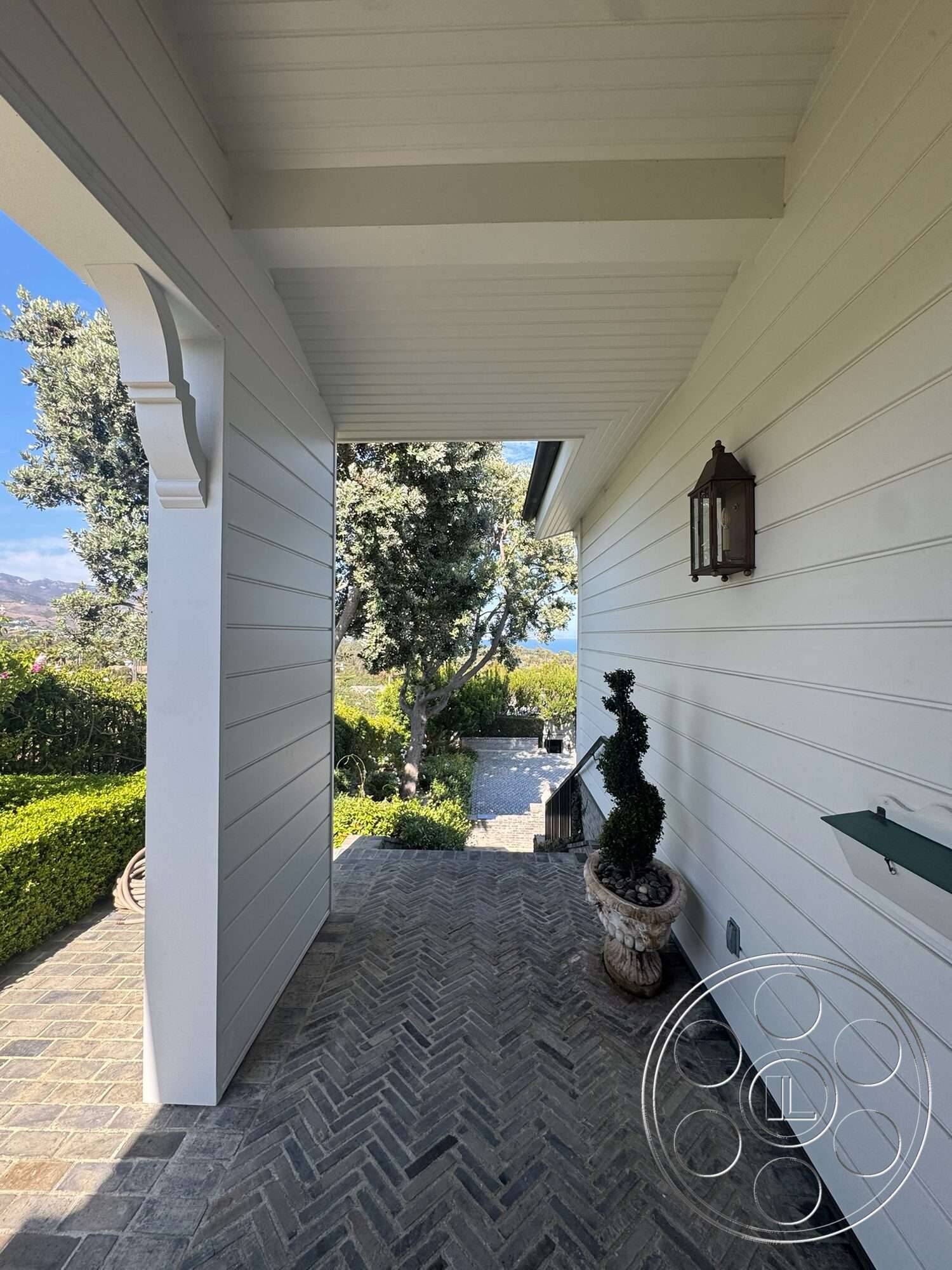 Americana 113 - exterior, surrounding greenery, wooden ceiling beams, white shiplap walls, arched entryway, clear sky view, landscaped garden area, decorative lantern light fixture, herringbone brick walkway, topiary plants in planters, stone steps leading up