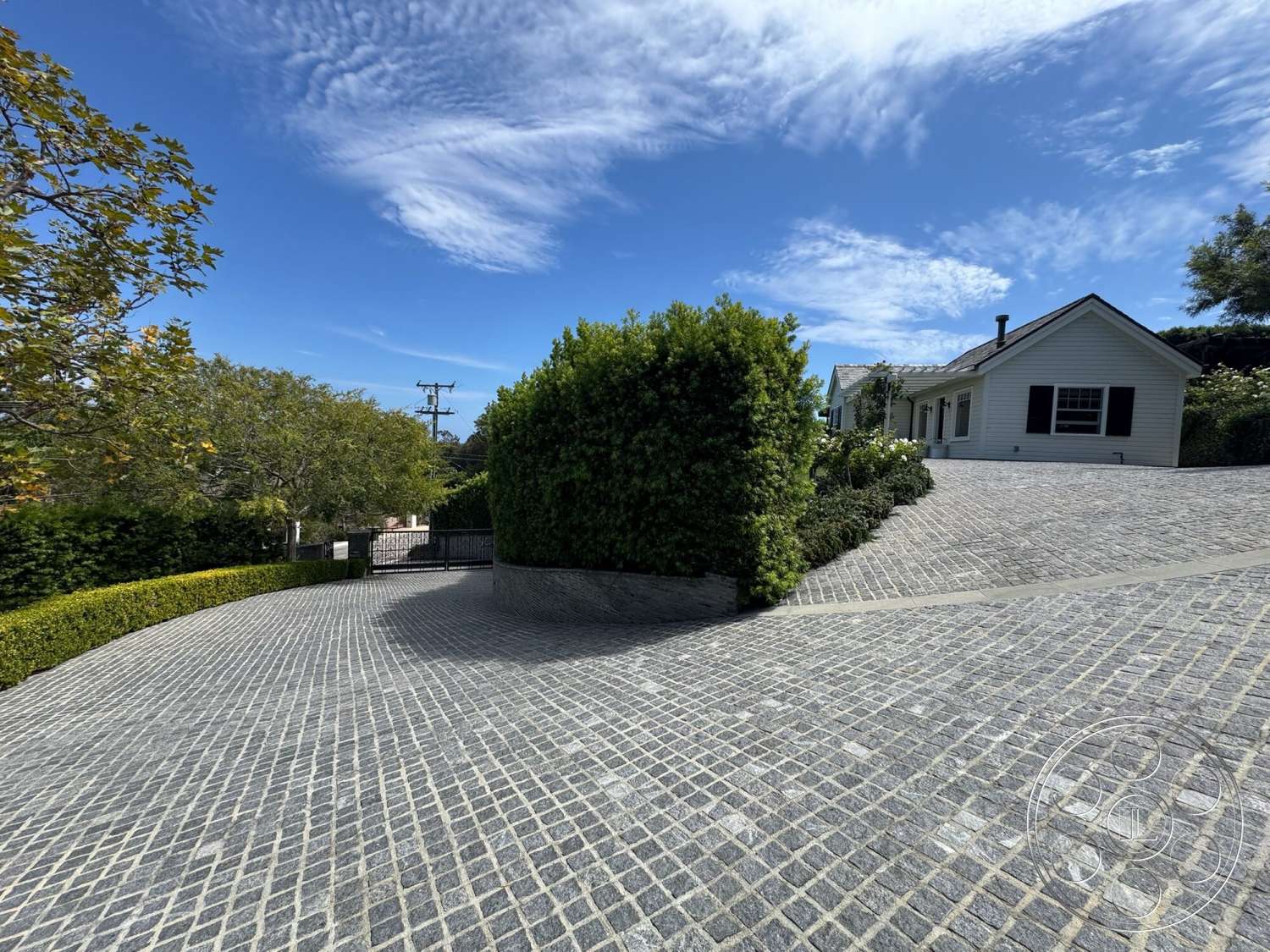 Americana 113 - curved driveway, hedge privacy screen, stone retaining wall, black window shutters, two-story home, landscaped garden area, tree-lined setting, paver stone driveway paving, white clapboard exterior, asphalt roof shingles