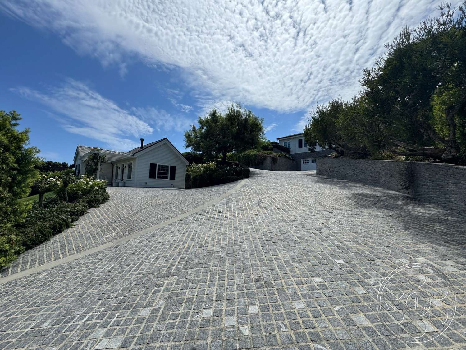 Americana 113 - exterior, planted shrubs, blue sky, sloped driveway, urban residential setting, cobblestone driveway, landscaped garden beds, stone retaining walls, textured paving stones, white siding house, dark window shutters, concrete path border
