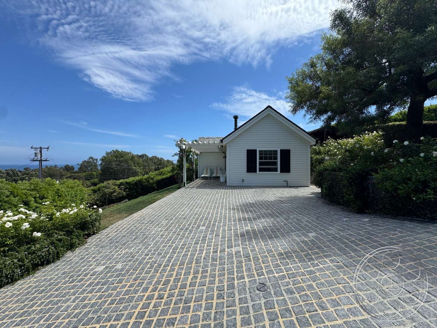 Americana 113 - exterior, paver driveway, sunlight, flowering shrubs, ocean view, black shutters, blue sky, landscape garden, gable roof, green hills, herringbone paver pattern, white wooden house