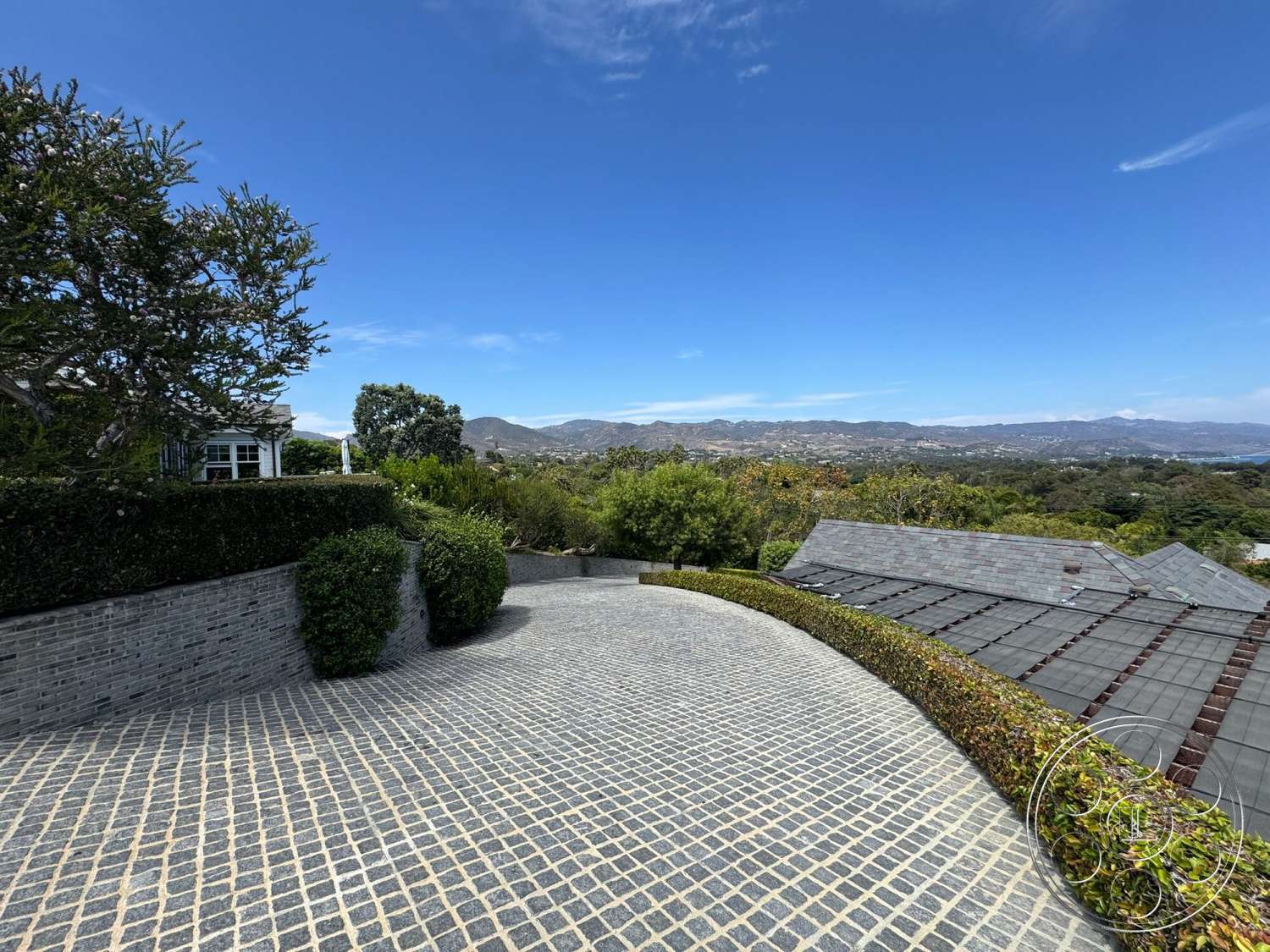 Americana 113 - exterior, lush greenery, cobblestone driveway, shingle roof, brick retaining wall, suburban hillside location, panoramic mountain view, landscaped hedge borders
