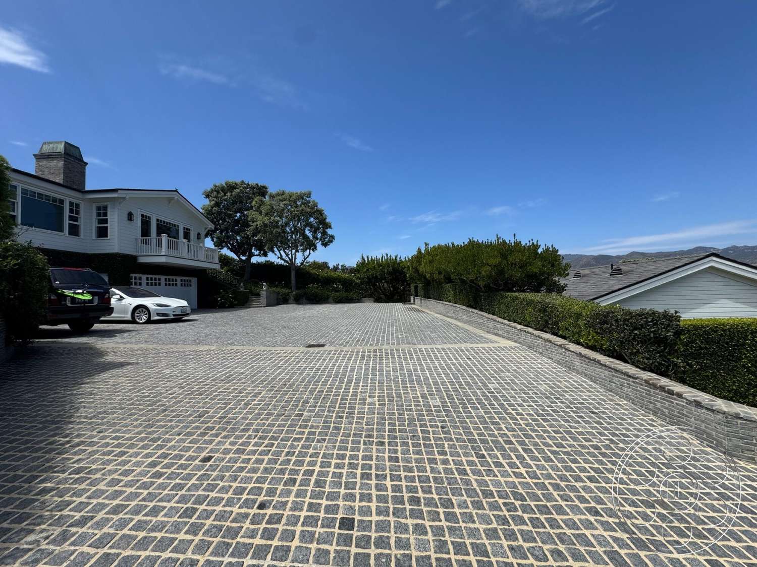 Americana 113 - paver driveway, gabled roof, clear blue sky, balcony with railings, urban residential, hilltop setting, landscaped hedges, two-story home, exterior siding, front yard greenery, cobblestone driveway surface
