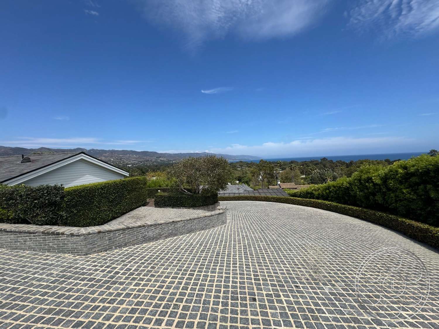 Americana 113 - exterior, surrounding hillside, single-story home, curved driveway, clear blue sky, cobblestone driveway, landscaped hedges, gentle slope, modern residential setting, distant ocean view