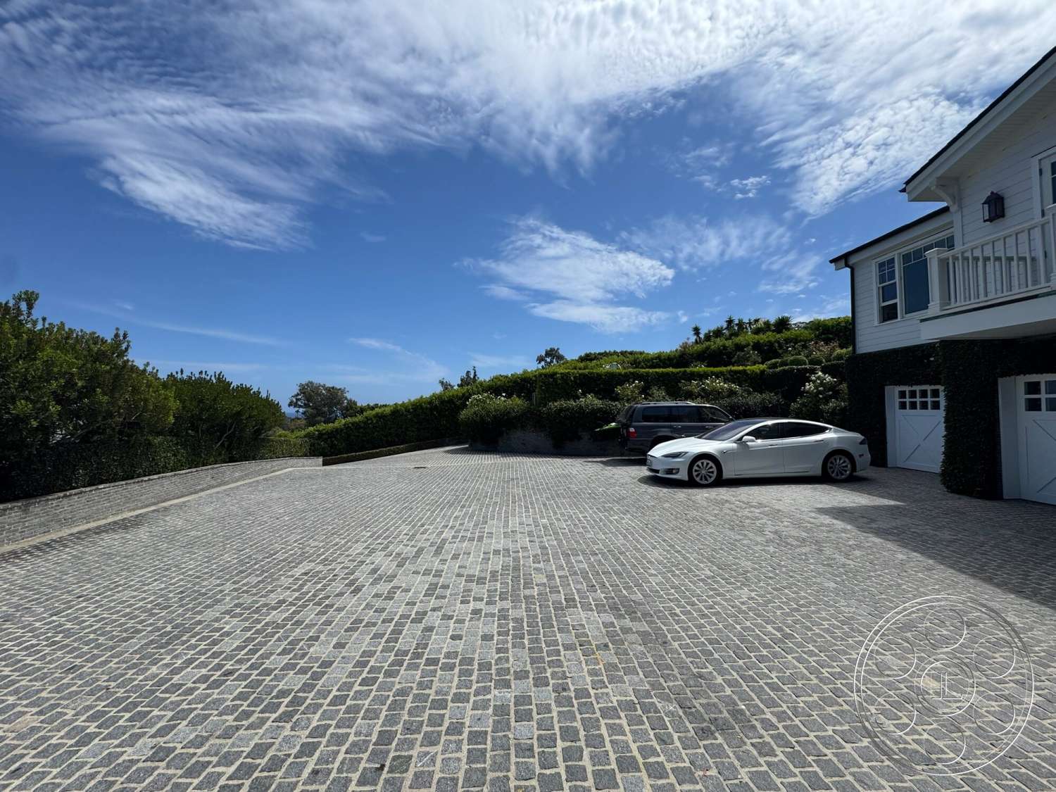 Americana 113 - large windows, exterior, white wooden siding, balcony with railing, gabled roof, clear blue sky, lush green hedges, cobblestone driveway, open parking area, overhanging eaves, two-story home, landscaped garden area, double garage doors