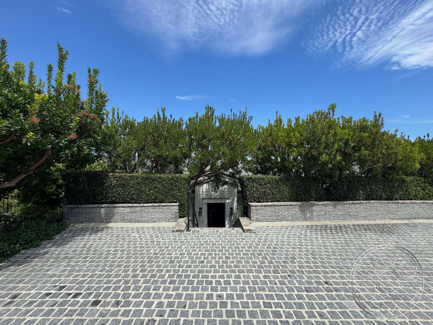 Americana 113 - exterior, modern architecture, minimalist aesthetic, urban setting, brick accent wall, symmetrical design, clear blue sky, greenery backdrop, cobblestone driveway, landscaped garden area, hedge boundary wall, large entrance steps