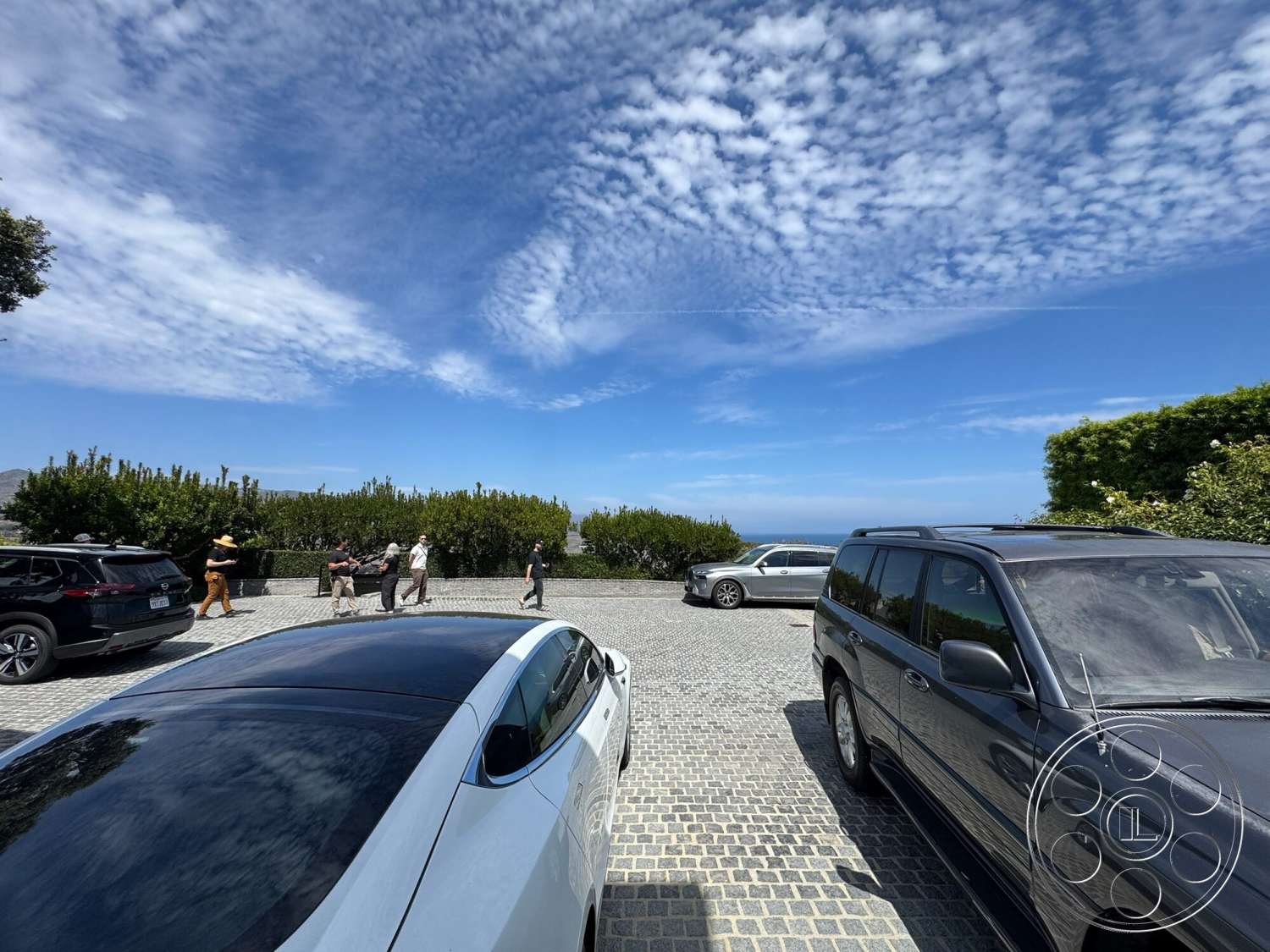 Americana 113 - exterior, ocean view, blue sky, cobblestone driveway, landscaped garden beds, outdoor gathering area, variety of parked vehicles, trees along the perimeter