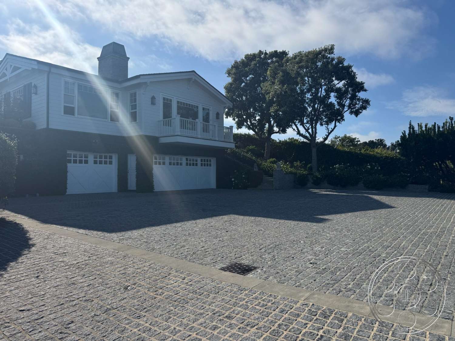 Americana 113 - exterior, landscaped garden, gravel pathway, white wood siding, balcony with railing, black accent wall, dormer roof, cobblestone driveway, large front porch, trees in background, decorative outdoor lanterns, double garage doors, raised two-story home