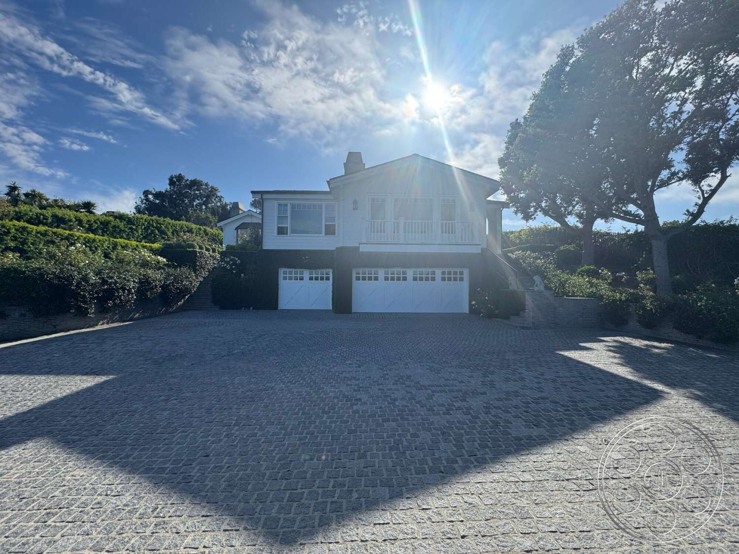 Americana 113 - exterior, mountain view, green hedges, white wood siding, gabled roof, garage doors, hillside location, cobblestone driveway, two-story home, landscaped garden beds, wooden balcony railing, stone driveway paving, sunshine lighting