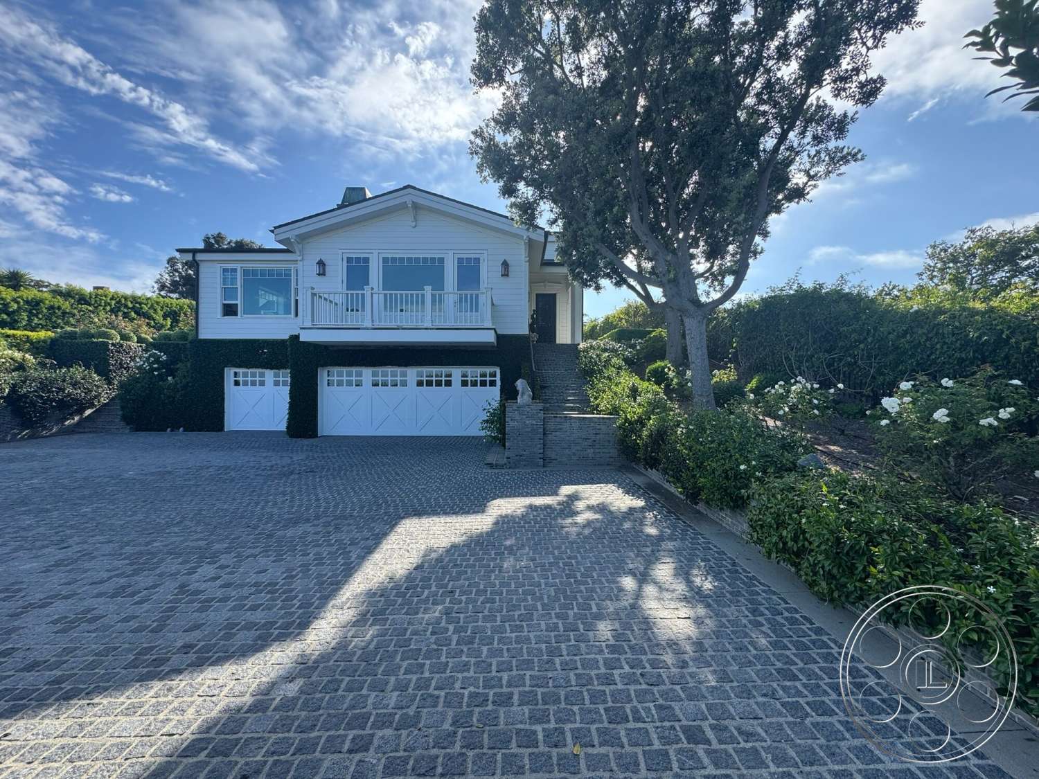 Americana 113 - exterior, landscaped garden, shingle siding, flowering shrubs, gabled roof, boxwood hedges, two-story home, white painted walls, brick driveway pavers, front porch with railing, large leafy tree, round-arched windows, cobblestone paving, pathway steps leading to entrance