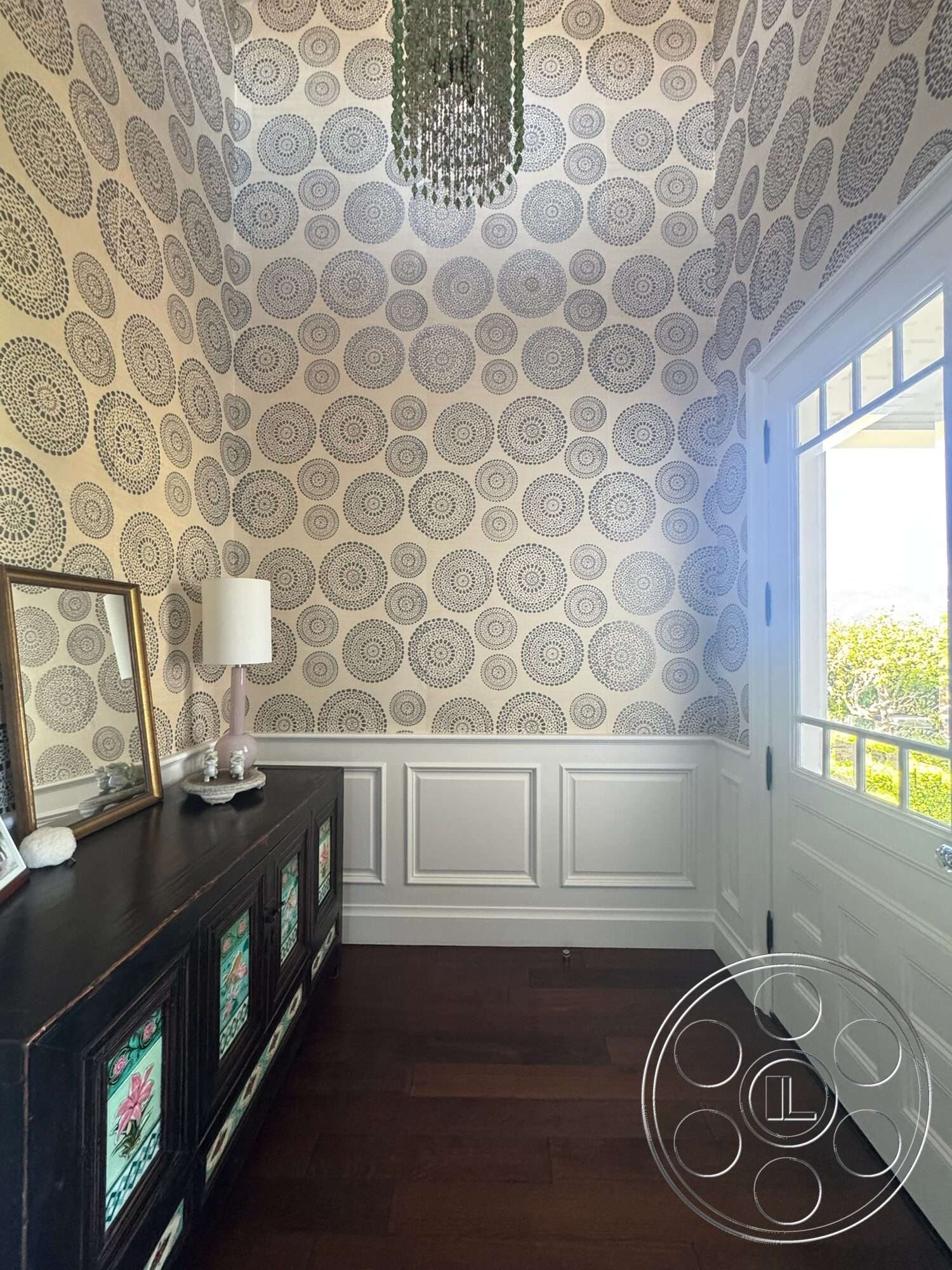 Americana 113 - arched doorway, white wainscoting wall treatment, textured wallpaper with circular patterns, dark hardwood entryway floor, glass chandelier fixture, black wooden console table with stained glass accents, framed mirror on console, natural light from entry door with divided glass panels