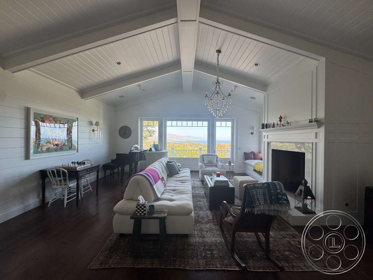 Americana 113 - textured area rug, white shiplap walls, large floor-to-ceiling windows, vaulted ceiling with exposed wood beams, chandelier lighting fixture, dark hardwood living room floors, white built-in fireplace surround, seating area with upholstered sofa, wooden dining table with white chairs, window seat with cushions, landscape view from windows
