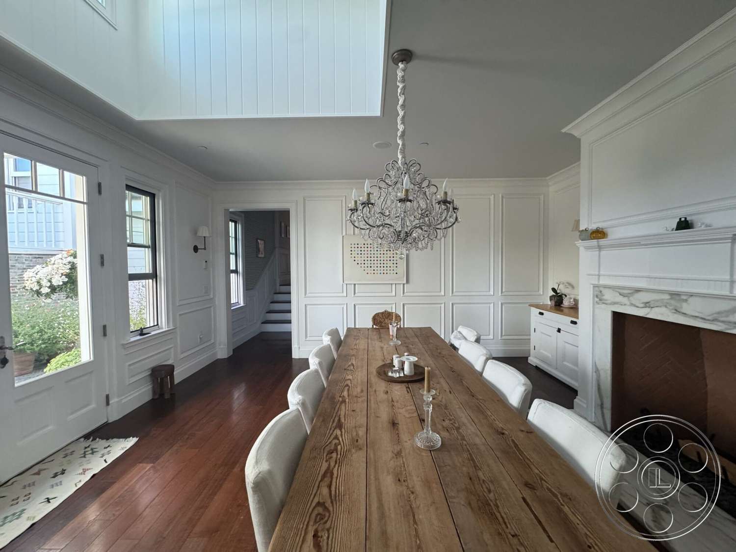 Americana 113 - neutral color palette, open floor plan, wood dining table, textured plaster ceiling, beaded chandelier, marble fireplace surround, hardwood dining room floors, white paneled walls, white kitchen cabinetry, white wainscoting detail, large windows with black trim, rectangular dining room layout