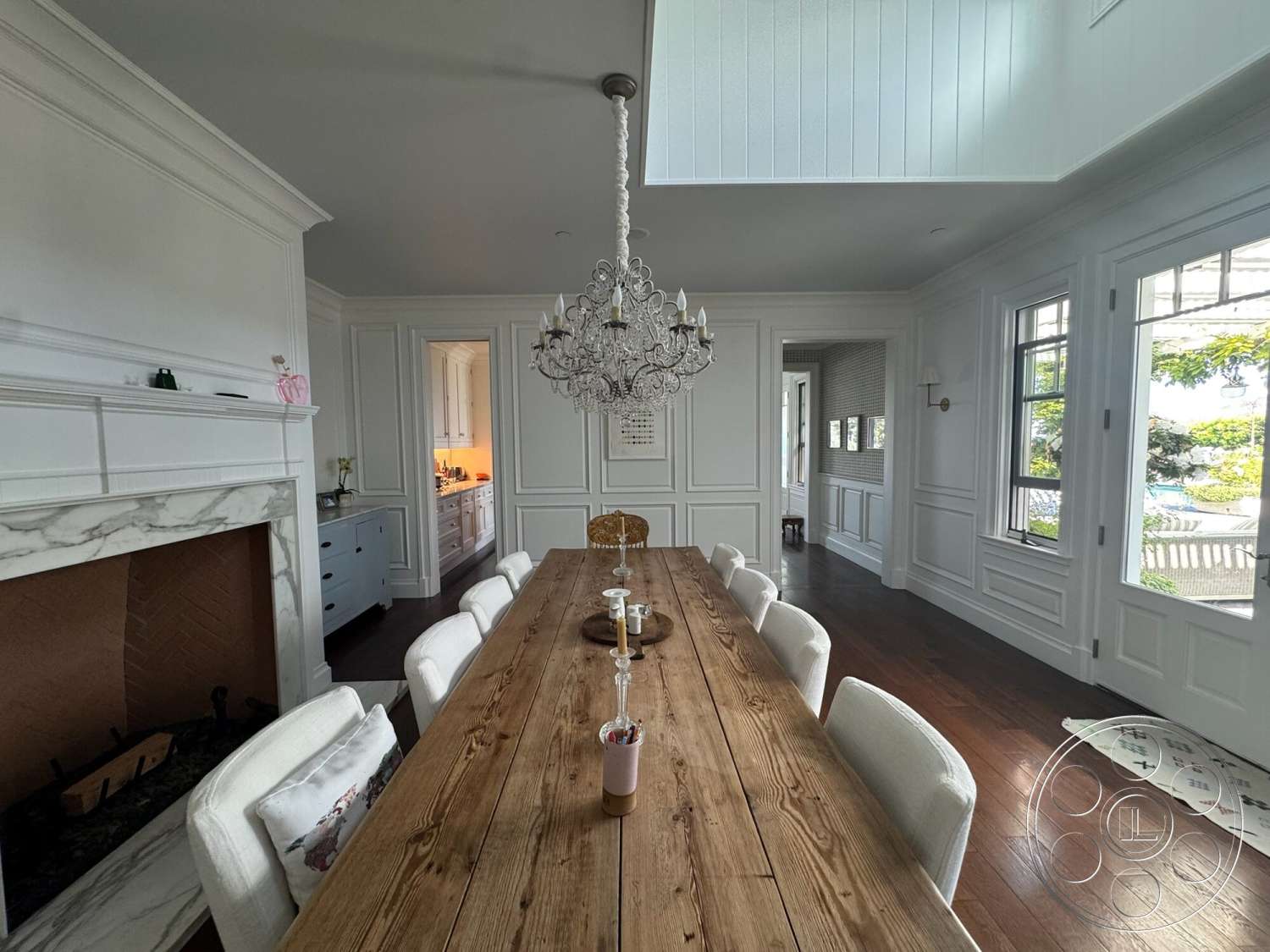 Americana 113 - open concept layout, upholstered dining chairs, floor-to-ceiling windows, chandelier lighting, arched doorway, long wooden dining table, built-in kitchen cabinets, marble fireplace surround, hardwood dining room floors, white wainscoting wall treatment, paneled ceiling