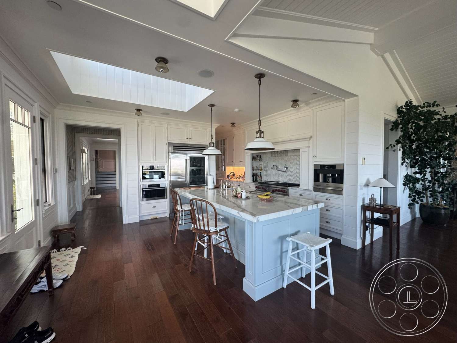 Americana 113 - stainless steel appliances, floor-to-ceiling windows, modern rustic decor, white shiplap walls, butler's pantry, pendant lighting fixtures, exposed wood ceiling beams, marble kitchen island countertop, open layout kitchen, hardwood kitchen floors, skylight ceiling, large indoor plant, raised panel kitchen cabinetry, white mosaic tile backsplash, island seating area, farmhouse style kitchen