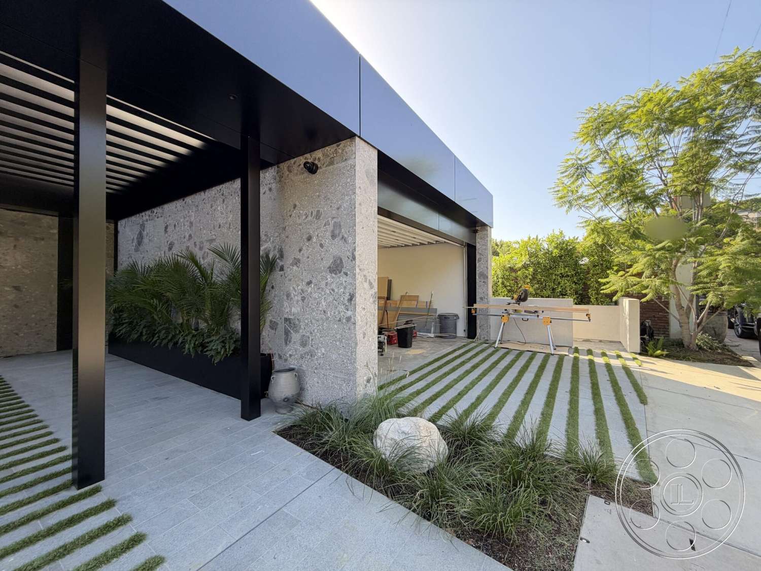 Modern 532 - exterior, concrete driveway, black metal roof, paver walkway, minimalist architectural style, horizontal wood slat overhang, grey stone cladding wall, striped grass landscaping, planter box with greenery, large boulders in landscaping