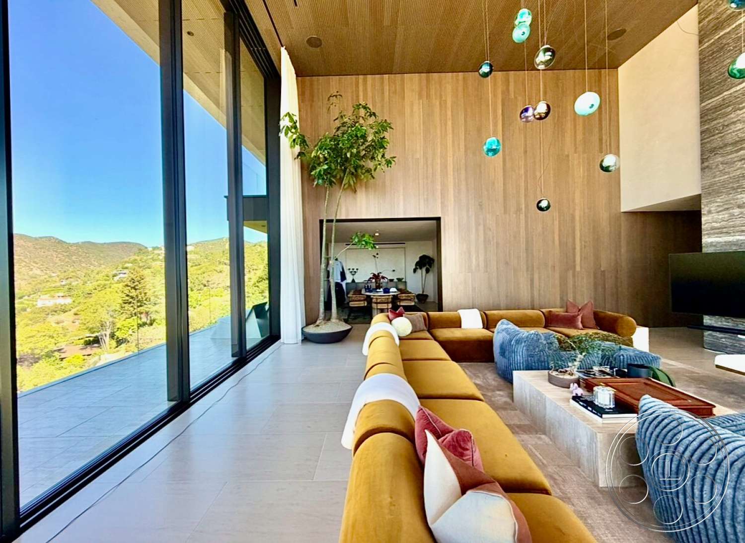 CK Mansion - open floor plan, floor-to-ceiling windows, textured throw pillows, mountain view, wood accent wall, concrete coffee table, contemporary furniture design, indoor potted plant, neutral tile flooring, light wood ceiling panels, sectional sofa with mustard upholstery, blue ribbed accent chairs