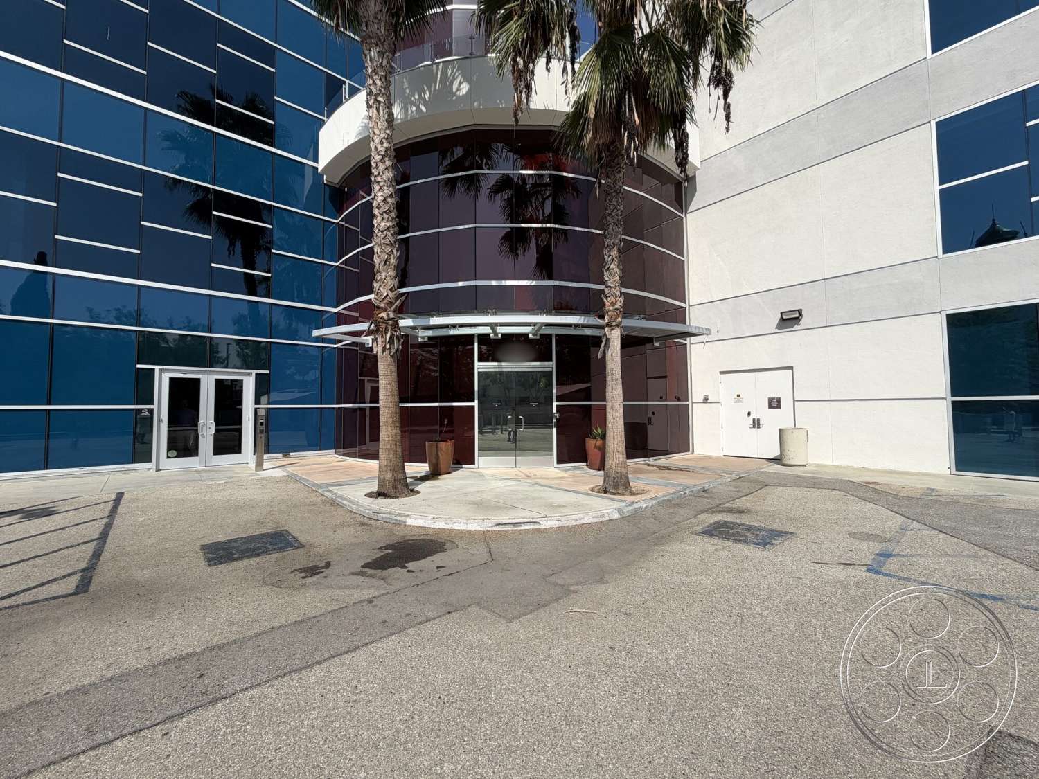 Office 130 - exterior, glass facade, modern architectural style, light-colored stucco walls, concrete walkway, large floor-to-ceiling windows, paved parking area, metal frame structure, planted palm trees, curved glass entrance canopy