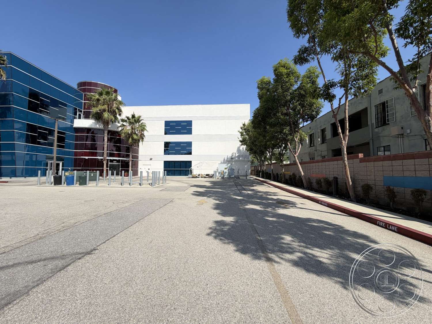 Office 130 - exterior, urban setting, glass façade, paved parking lot, concrete sidewalk, modern commercial architecture, gray concrete surface, painted concrete walls, blue glass panels, red curved structure, landscaped area with palm trees, metal barriers