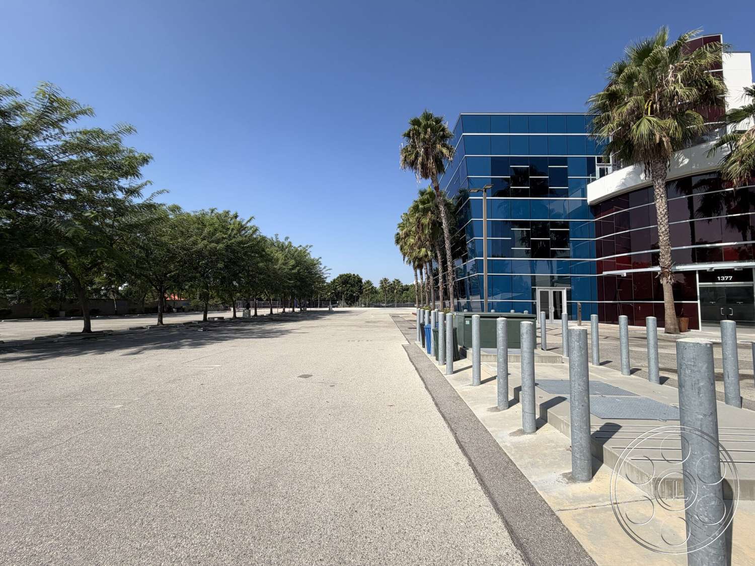 Office 130 - exterior, concrete pavement, clear blue sky, asphalt parking lot, metal bollards, modern commercial building, landscaped area with palm trees, blue glass facade, red glass accents