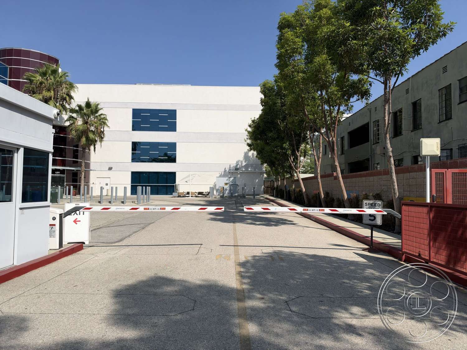 Office 130 - exterior, urban setting, concrete driveway, concrete walkways, metal security gate, modern industrial architecture, glass window panels, palm trees lining the entrance, white painted building facade, striped security gate, red accent walls, commercial property entrance
