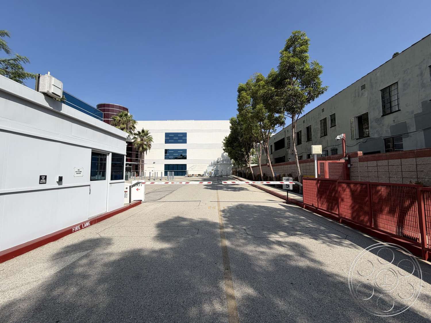 Office 130 - exterior, textured concrete walls, urban setting, glass windows, concrete driveway, clear blue sky, landscaped with palm trees, modern industrial building, exposed aggregate surface driveway, red metal gate, white paneled security building, linear pattern on facade, two-story industrial structure