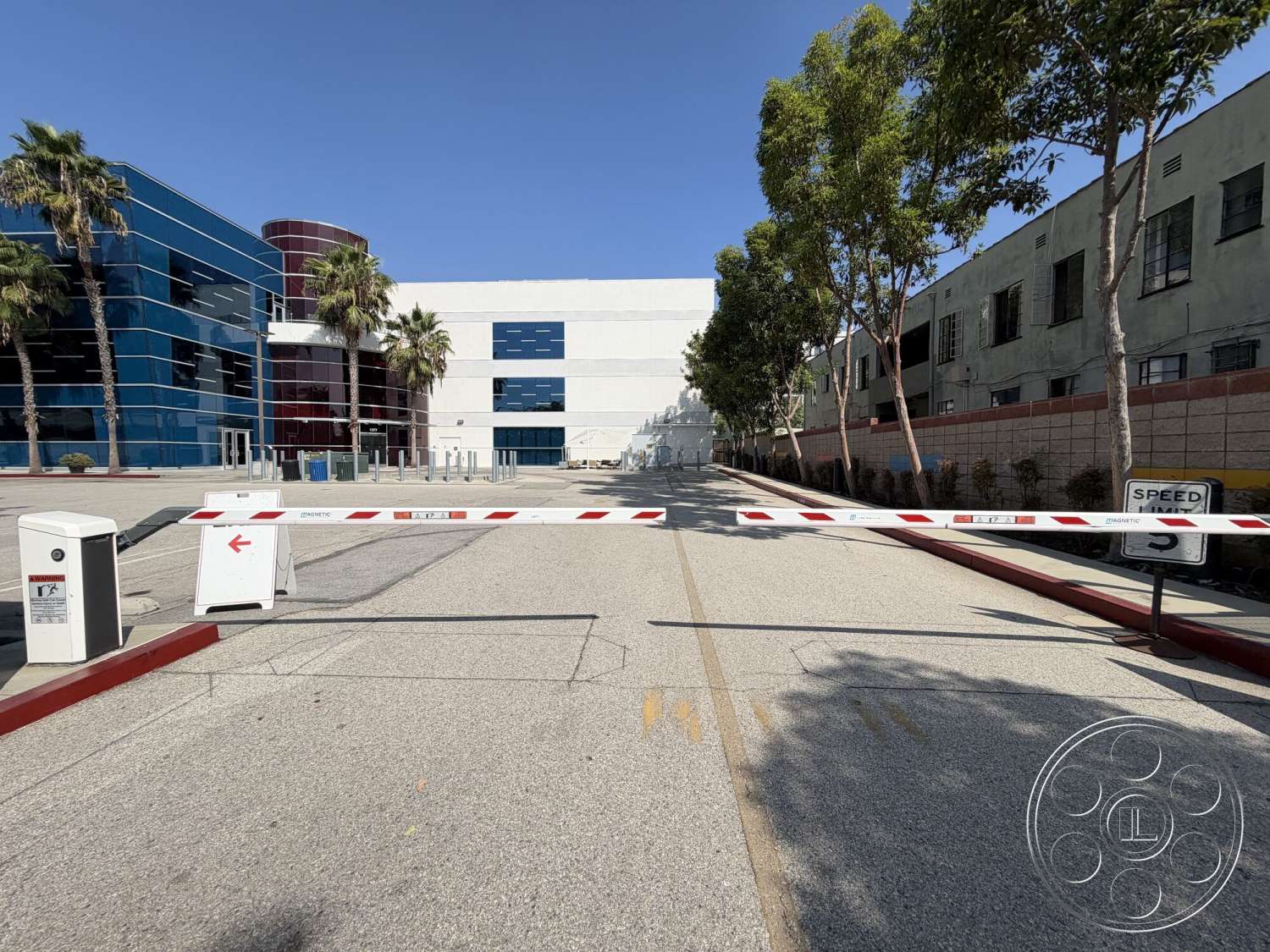 Office 130 - exterior, urban environment, modern architectural style, concrete parking lot, palm trees landscaping, striped road markings, glass building facade, blue glass panels, red cylindrical structure, security barrier gate, industrial building design, sidewalk pavers, concrete perimeter wall