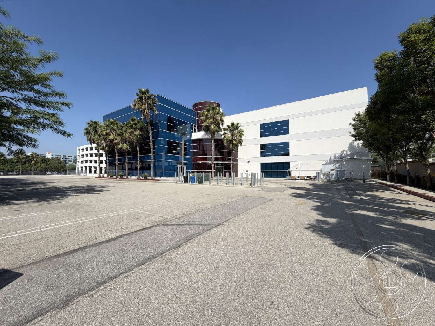Office 130 - exterior, white stucco walls, modern architectural style, large floor-to-ceiling windows, concrete parking lot, flat roof structure, palm trees landscaping, blue glass facade, circular red glass tower, horizontal window accents