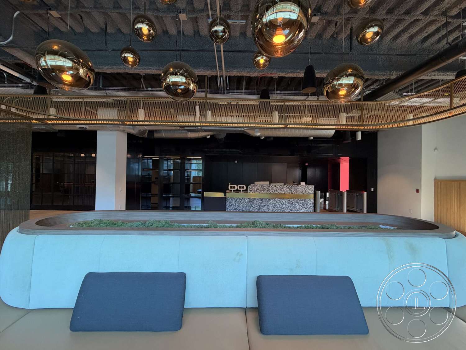 Office 130 - black cabinetry, open floor plan design, exposed ductwork ceiling, textured fabric sofa cushions, suspended gold pendant lighting, curved blue upholstered seating, wood accent trim, natural greenery feature, stone reception desk