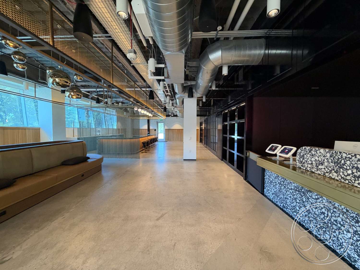Office 130 - neutral color palette, modern interior, polished concrete floors, floor-to-ceiling windows, suspended light fixtures, open office layout, glass partition walls, exposed concrete flooring, industrial style decor, metal ceiling beams, seating area with upholstered bench, contemporary workspace environment, wood panel walls, reception desk with stone front, elongated bar-height table