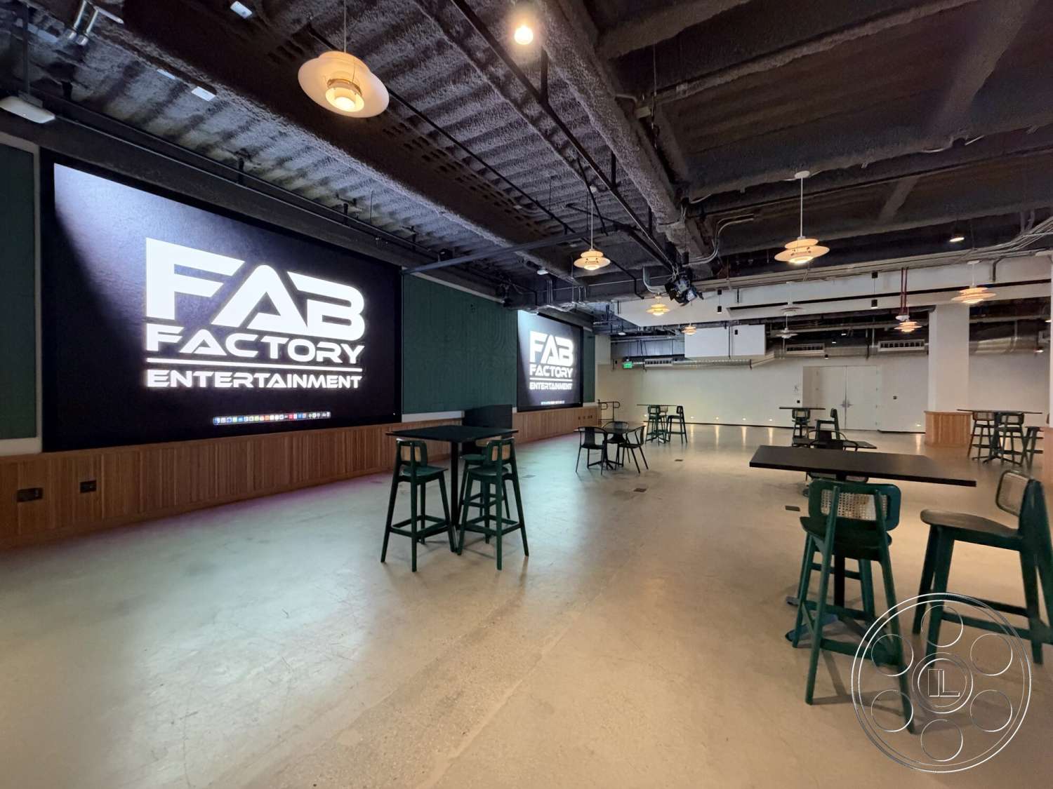 Office 130 - minimalist design, modern aesthetic, high-top seating, exposed concrete flooring, industrial-style lighting fixtures, rectangular tables, black ceiling with exposed ductwork, wood panel wall with logo, open entertainment space