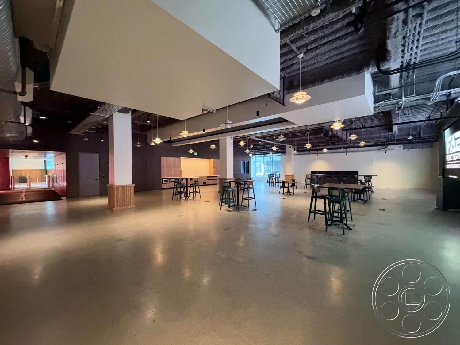 Office 130 - open floor plan, bright natural light, polished concrete floors, black metal chairs, rectangular layout, wood panel accent wall, exposed ductwork ceiling, industrial style lighting, glass door entry, pendant light fixtures, bar height tables