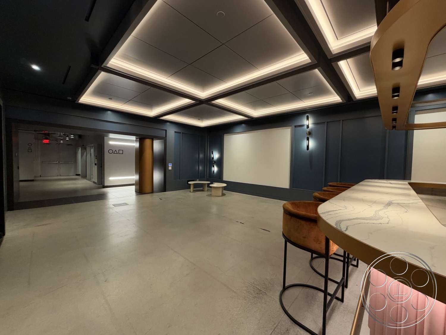 Office 130 - minimalist design, open floor plan, polished concrete flooring, marble countertop with veining, dark blue wall treatment, coffered ceiling with recessed lighting, gold accent bar counter, modern bar stools with brown upholstery, built-in wall lighting, elevator access with mirrored wall, multi-functional lounge area