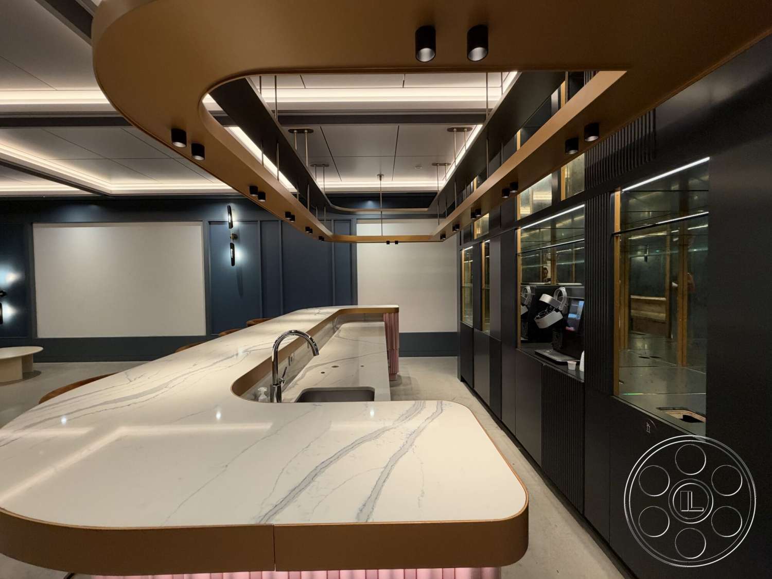 Office 130 - dark cabinetry, integrated appliances, smooth concrete flooring, modern kitchen island, gold accent lighting, open kitchen layout, marble kitchen countertop, minimalist kitchen design, linear ceiling lights, wood cabinet details, high-tech coffee station, blue feature wall, sleek kitchen fixtures